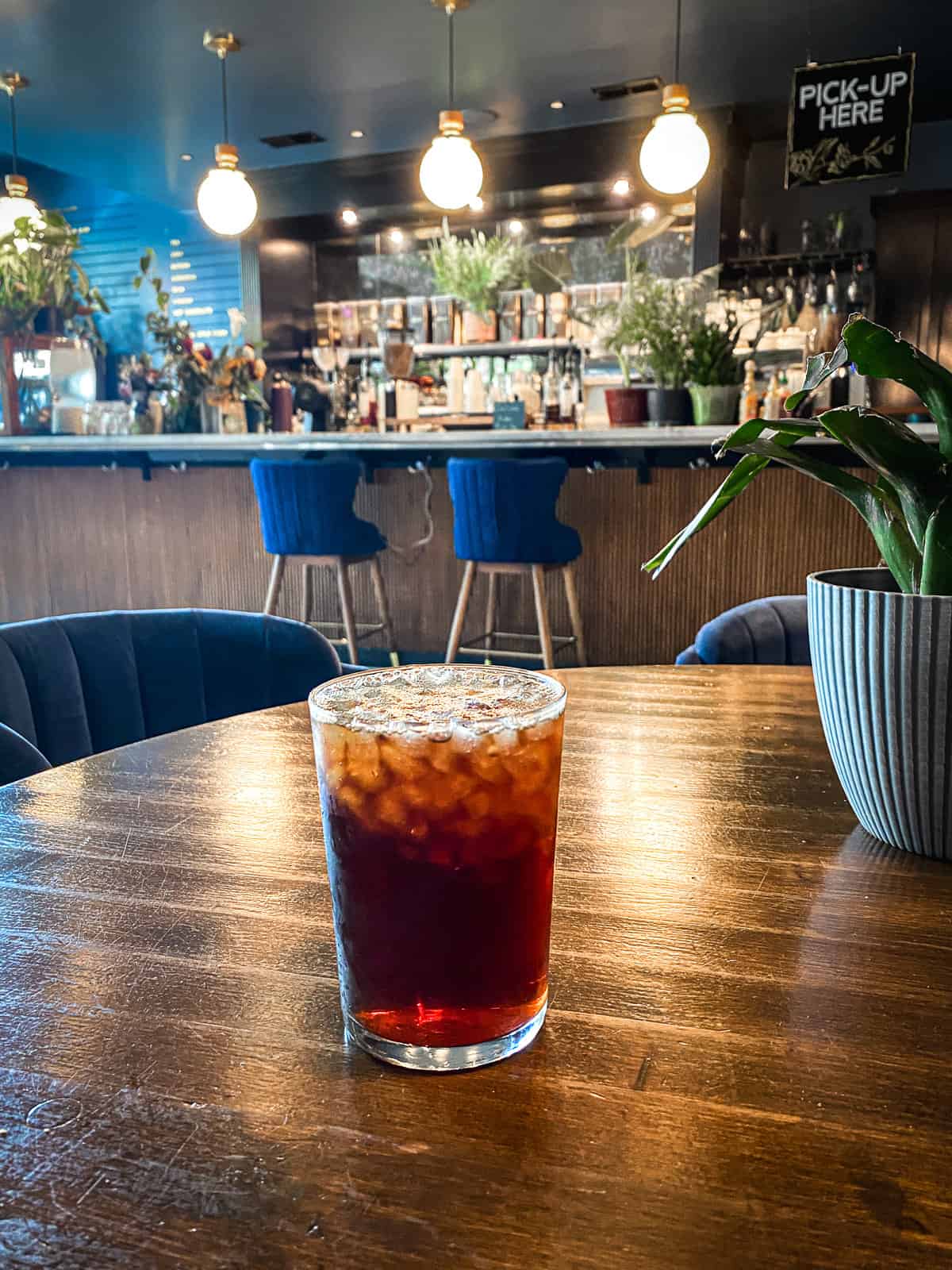 An iced coffee in a trendy coffee shop with dark blue walls, dark wood and lots of green plants.