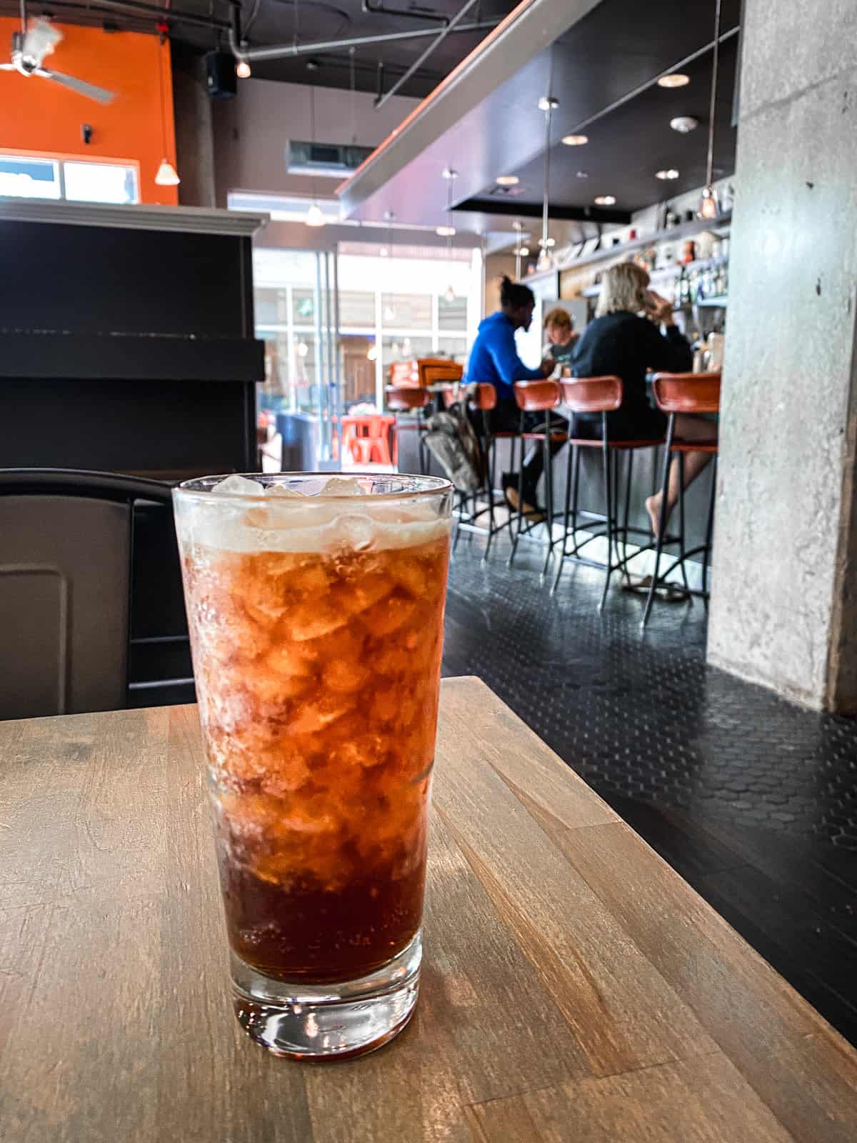 A large iced coffee on a table in a dark, industrial style coffee shop with people working at long tables