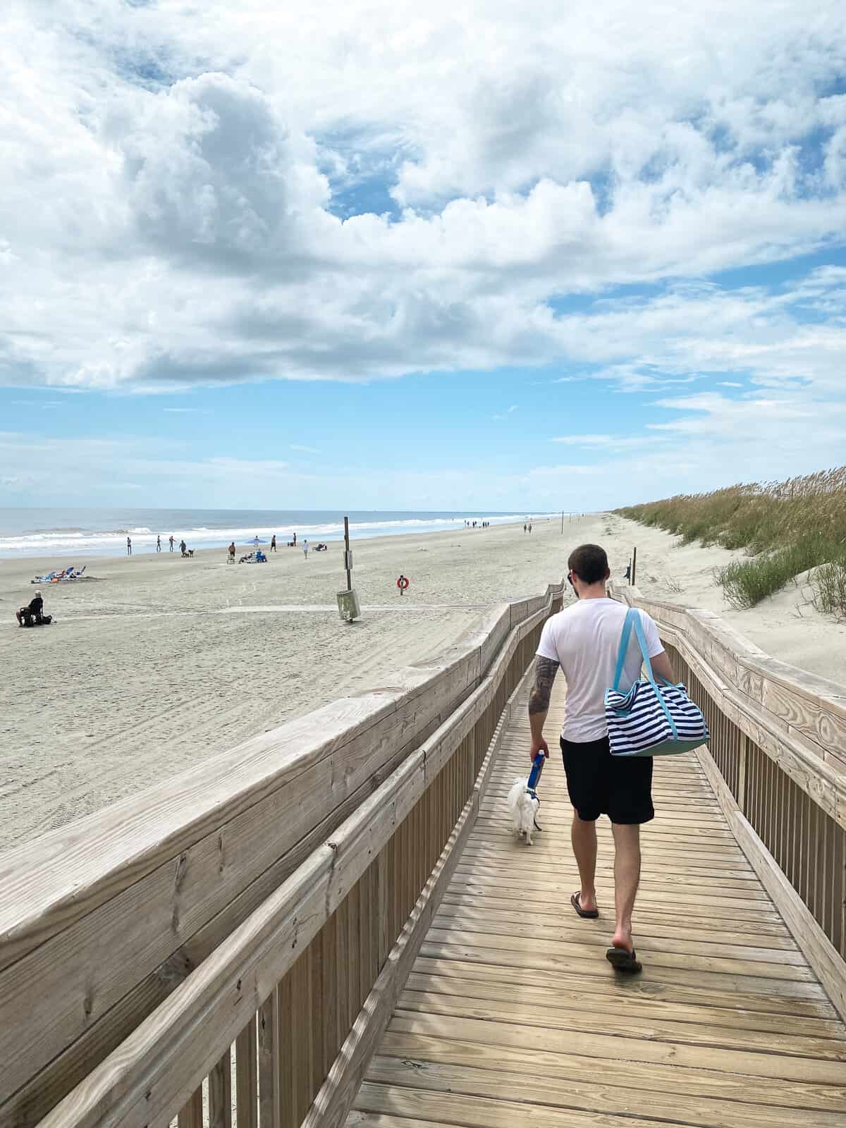 A man walking a small white dog on a sunny, beach boardwalk