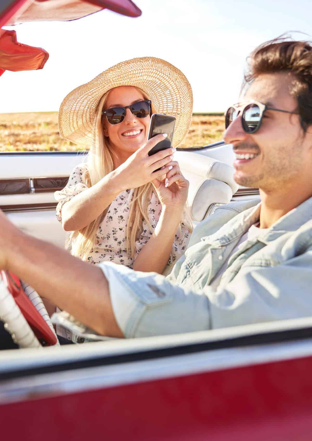 woman in a sun hat taking pictures of a man as he drives a convertible. 