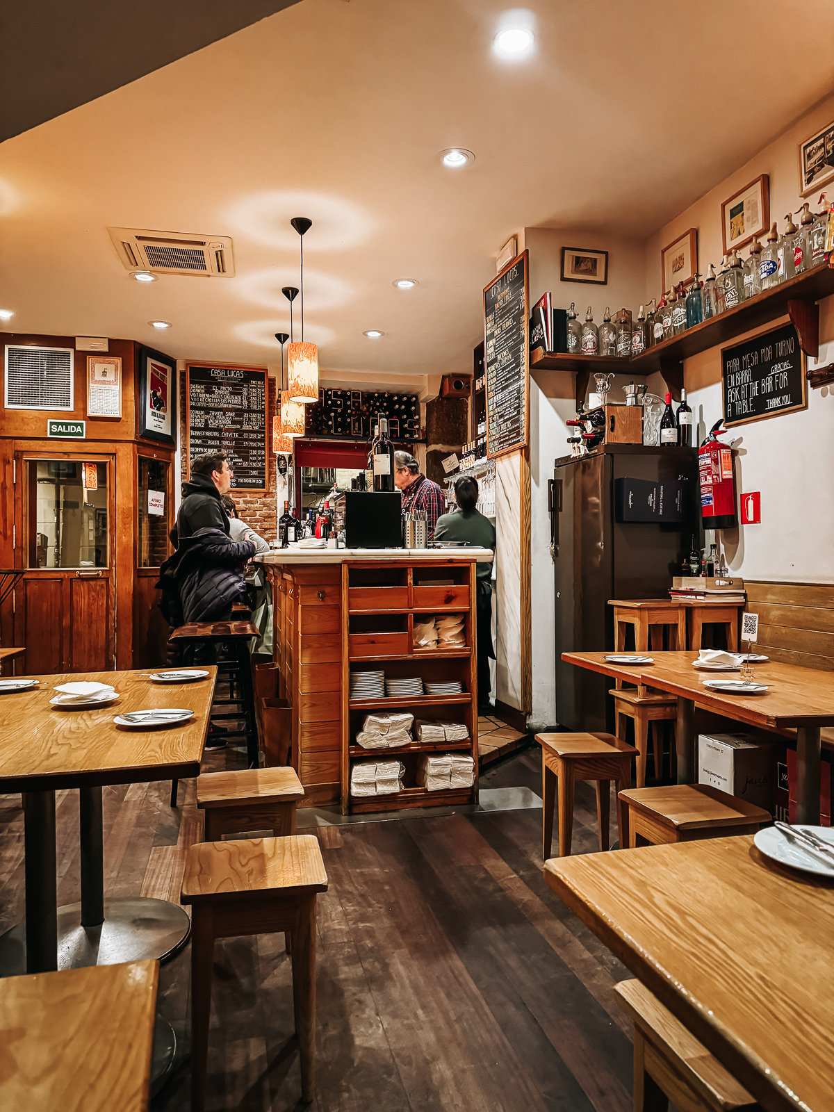 Cozy restaurant interior with wooden tables and chairs, patrons seated at the bar, and a menu board in the background, conveying a warm and inviting dining experience