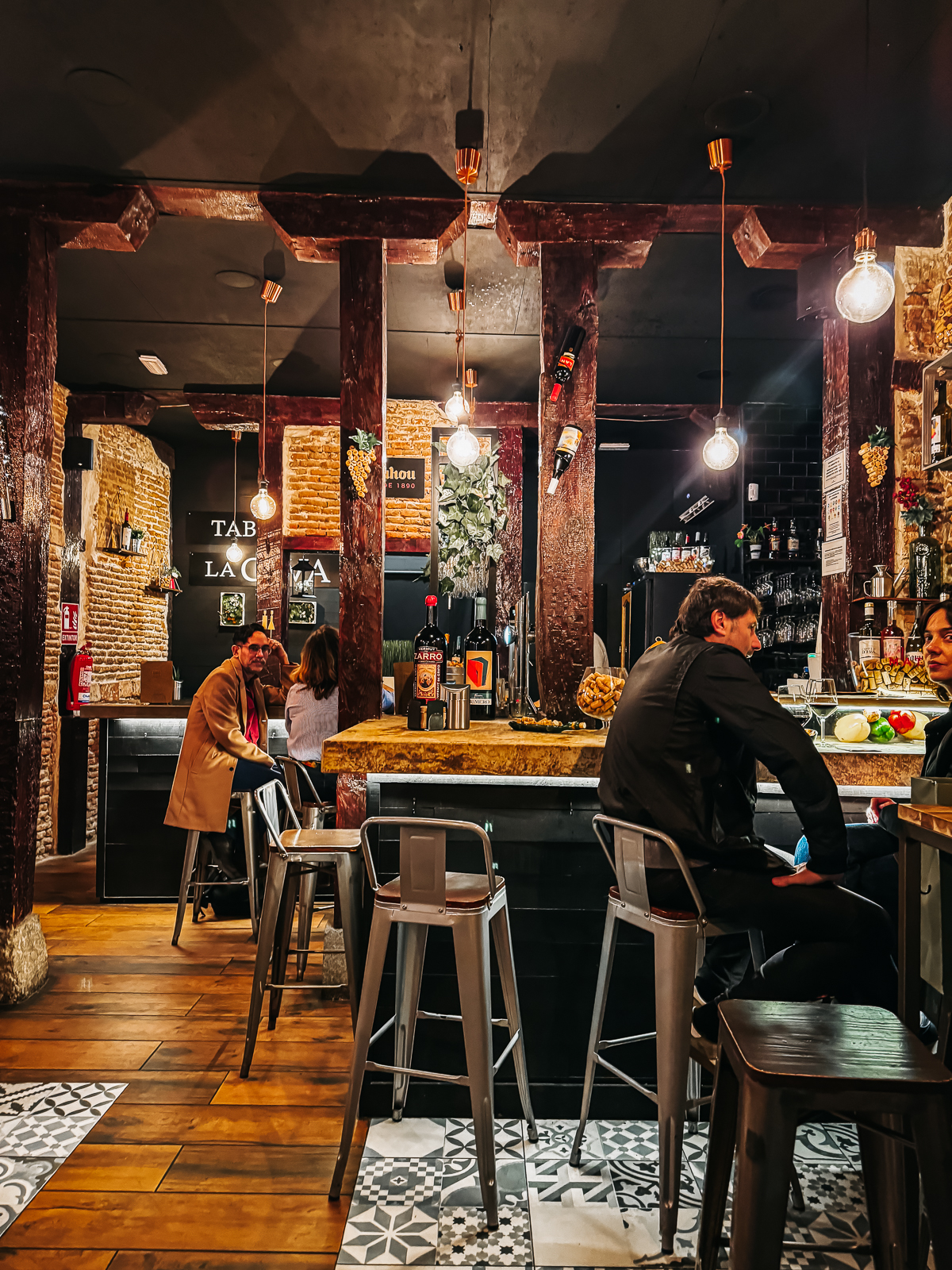Rustic bar interior with exposed brick walls and wooden beams, patrons seated at the bar, and decorative hanging light bulbs creating a warm, inviting ambiance.