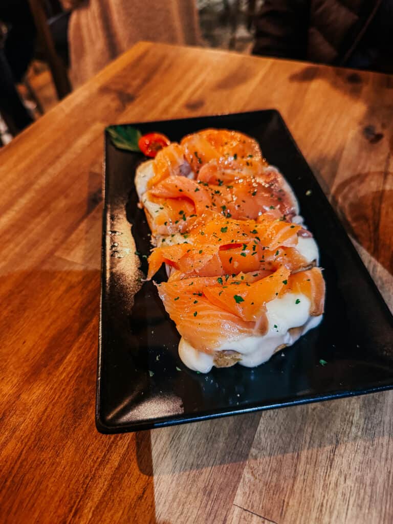 A gourmet dish presenting thinly sliced smoked salmon over a bed of cream cheese on toast, garnished with fresh herbs, on a sleek black rectangular plate.