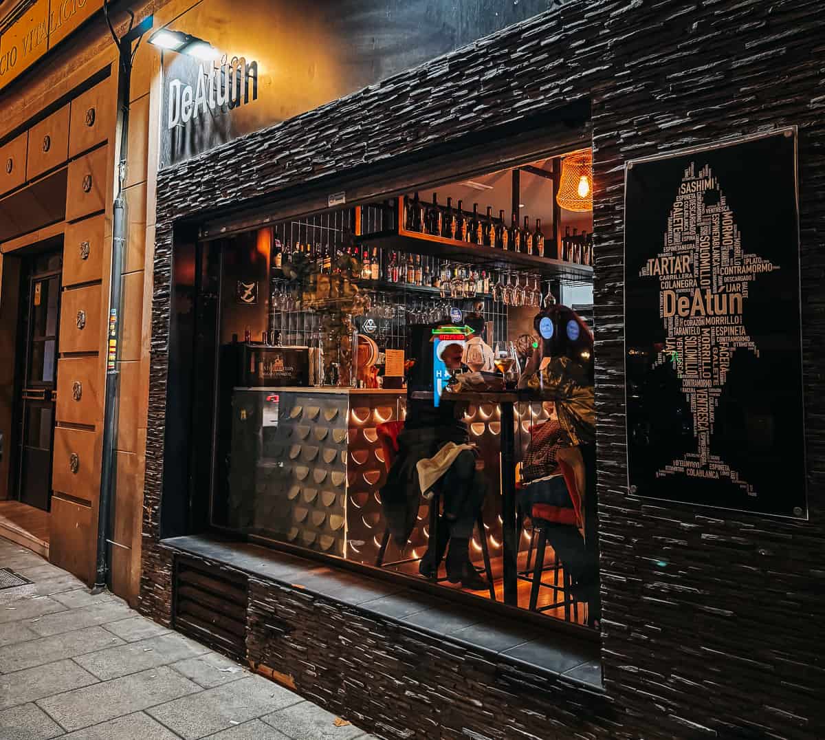 A stylish tapas bar with stone walls and a sign that reads "DeAtun" with patrons sitting and dining in the window