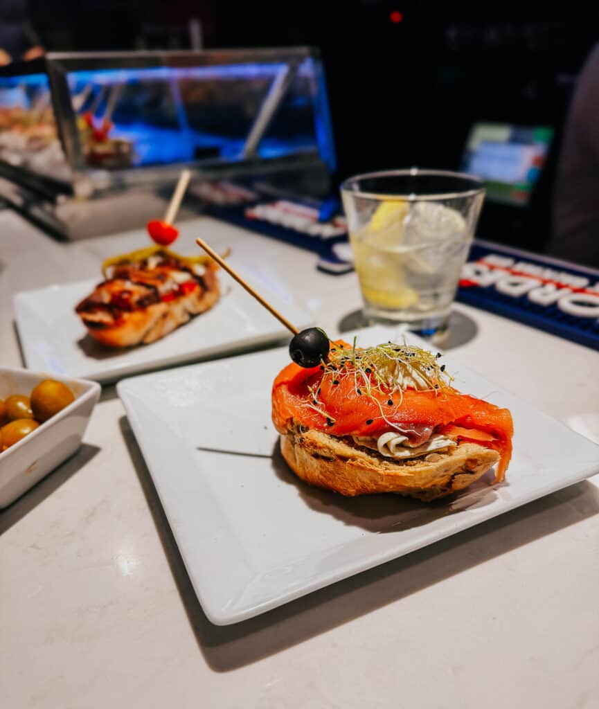 Elegant presentation of a tapa with smoked salmon and sprouts on crusty bread, accompanied by another tapa and a bowl of olives, set on a marble countertop.