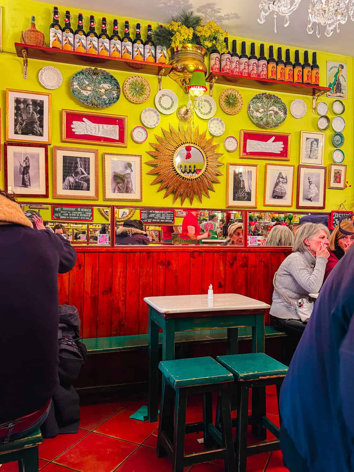 Vibrant bar wall decorated with eclectic artwork, plates, and bottles, under a green ceiling, creating a lively and colorful atmosphere.