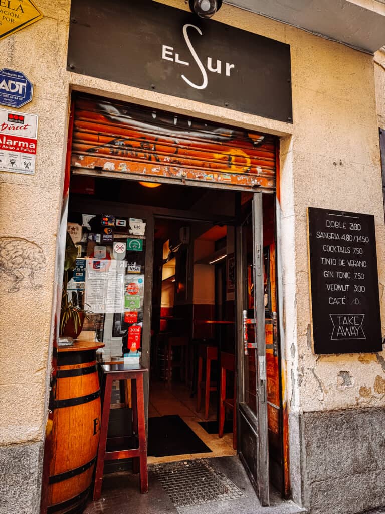 Close-up of the entrance to 'Taberna El Sur', showcasing a colorful shutter above the door, various stickers and signs, and a chalkboard with drink specials including 'Sangria' and 'Gin Tonic