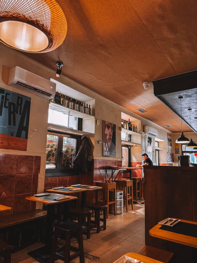 nterior of 'Taberna El Sur' with its warm, inviting atmosphere, featuring wooden tables and floor, movie posters on the wall, and a large woven lampshade overhead