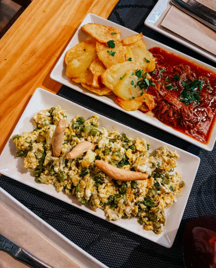 Delicious Spanish tapas on white plates at 'Taberna El Sur', including 'Patatas Bravas', 'Revuelto de Bacalao', ropa vieja, nd a bread slice, all served on a wooden table with cutlery.