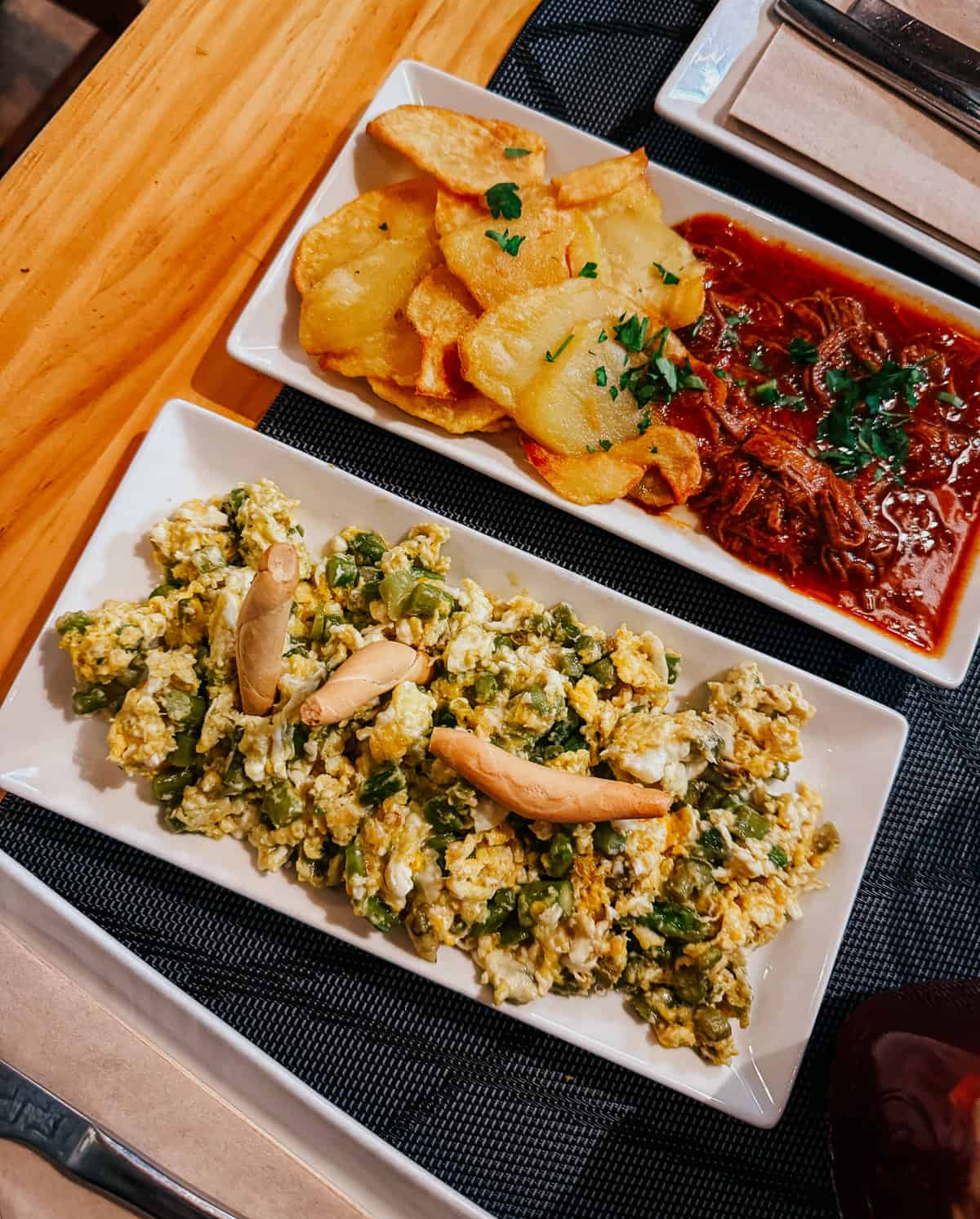 Delicious Spanish tapas on white plates at 'Taberna El Sur', including 'Patatas Bravas', 'Revuelto de Bacalao', ropa vieja, nd a bread slice, all served on a wooden table with cutlery.