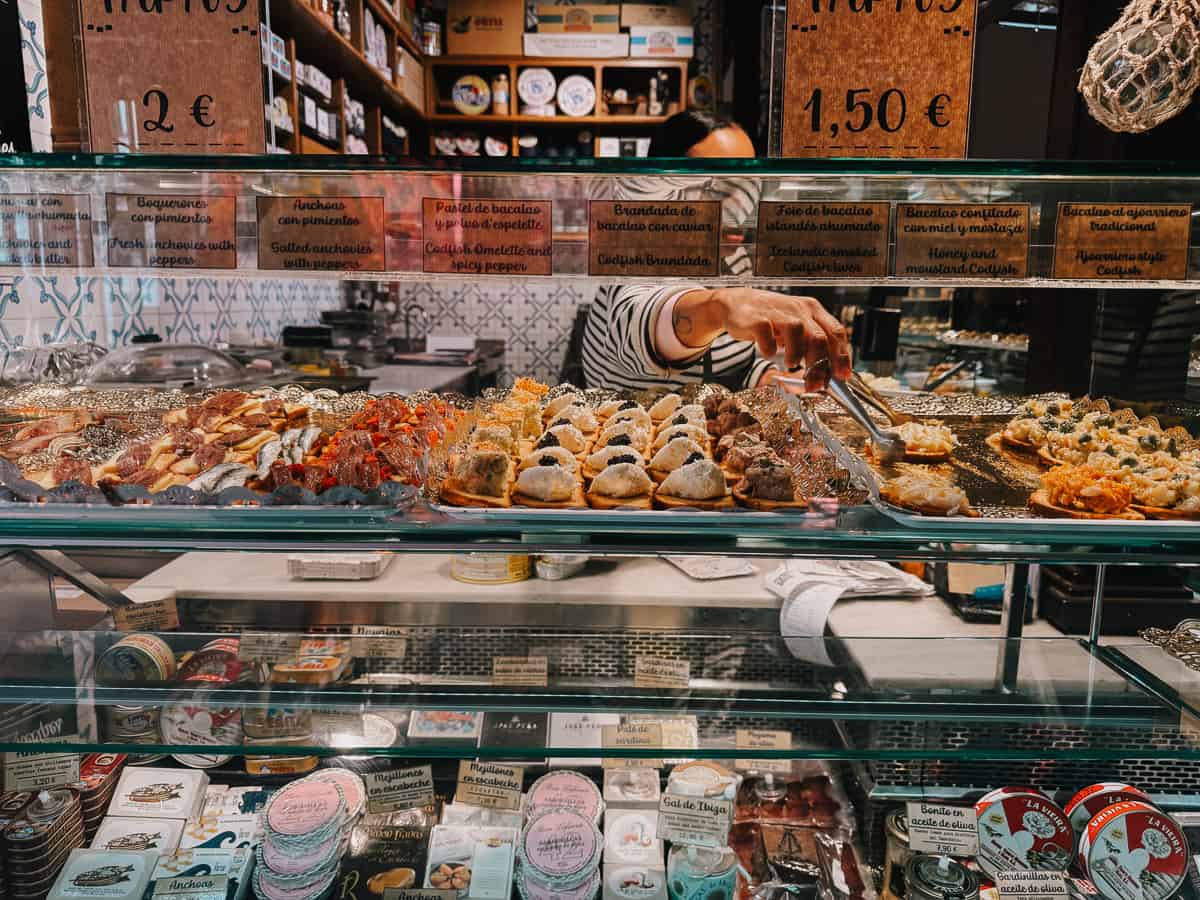 A vibrant display of Spanish tapas at a market stall, with a server arranging assorted delicacies like pastel de bacalao, boquerones, and anchoas. Prices are visible, offering a glimpse into the local culinary commerce.
