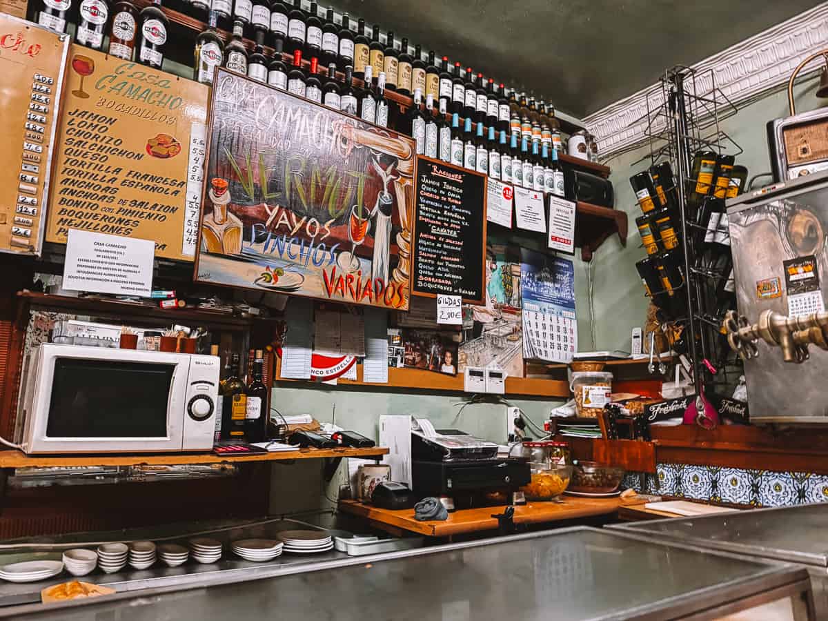 Interior of Casa Camacho showcasing a rustic bar with an array of tapas and vermouth offerings, surrounded by shelves filled with wine and spirits bottles, and a menu board listing various Spanish snacks