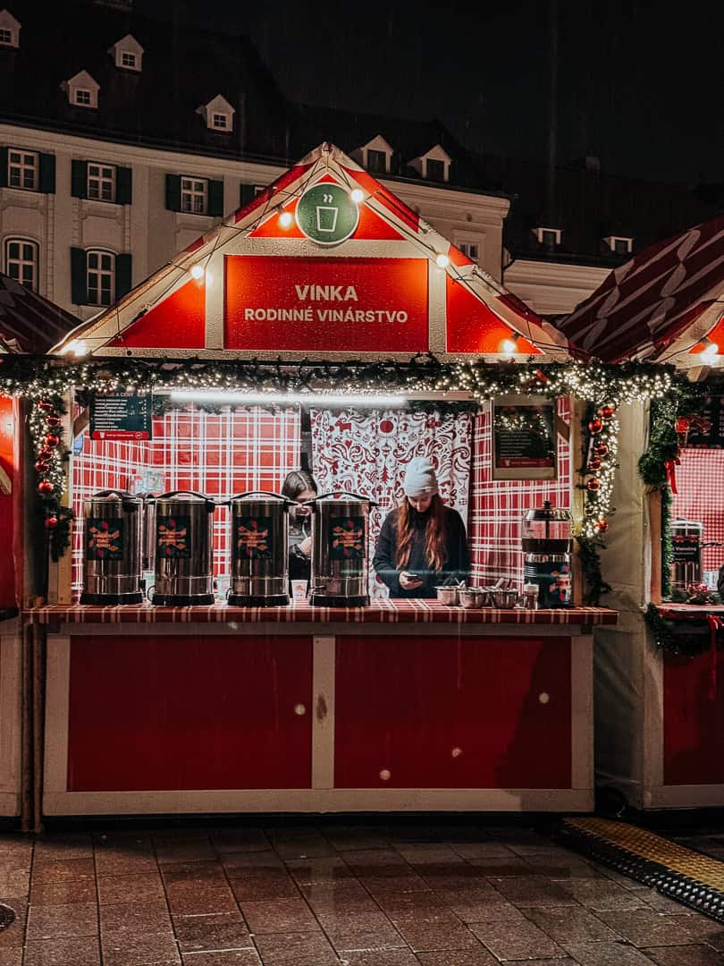 A vibrant holiday market stall at night with the sign "VINKA Rodinne Vinárstvo," decorated with festive lights and garlands, showcasing wine dispensers and a vendor interacting with a customer.