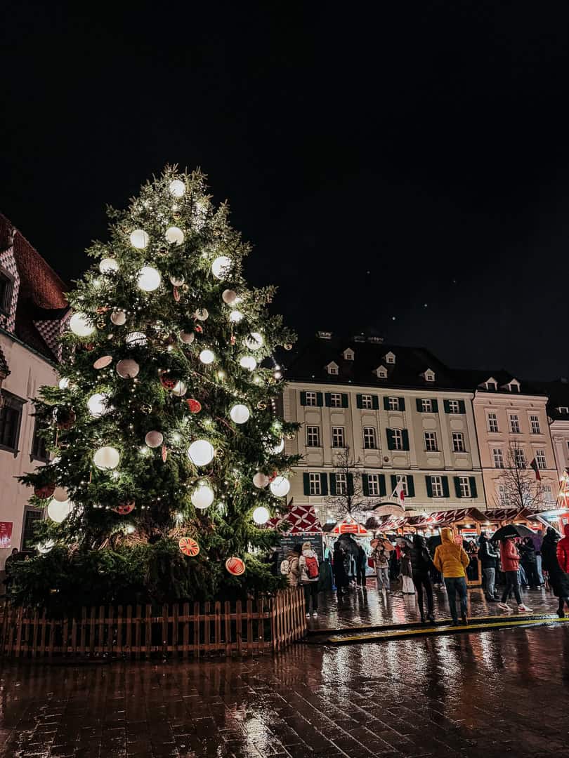 A beautifully lit Christmas tree standing tall in a night market, surrounded by bustling crowds of shoppers and festive red-and-white-striped market stalls.