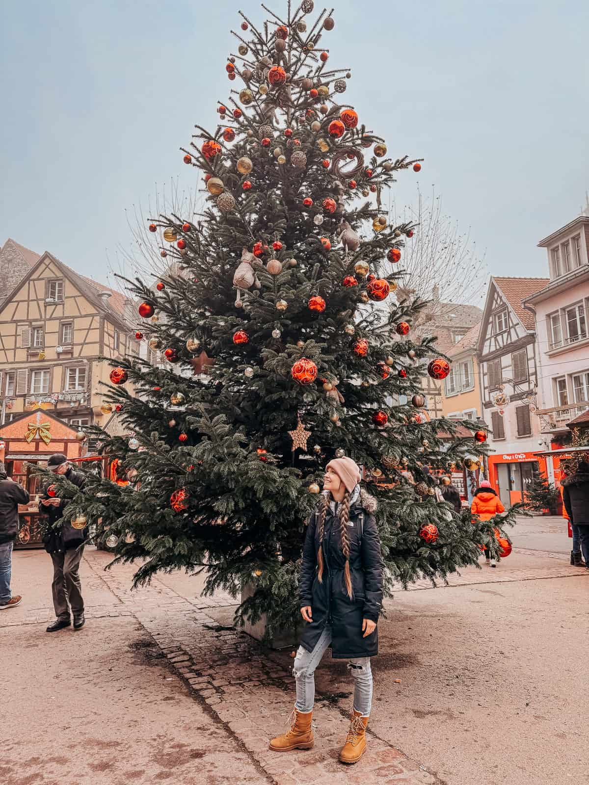 A person stands in awe before a towering Christmas tree beautifully decorated with red and gold ornaments in the festive streets of Colmar."