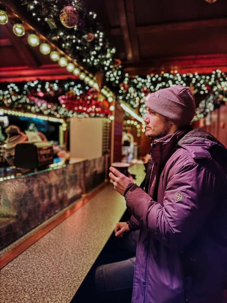 A man in a purple jacket enjoying a hot beverage at a Christmas market, with twinkling lights and holiday decorations enhancing the festive mood.