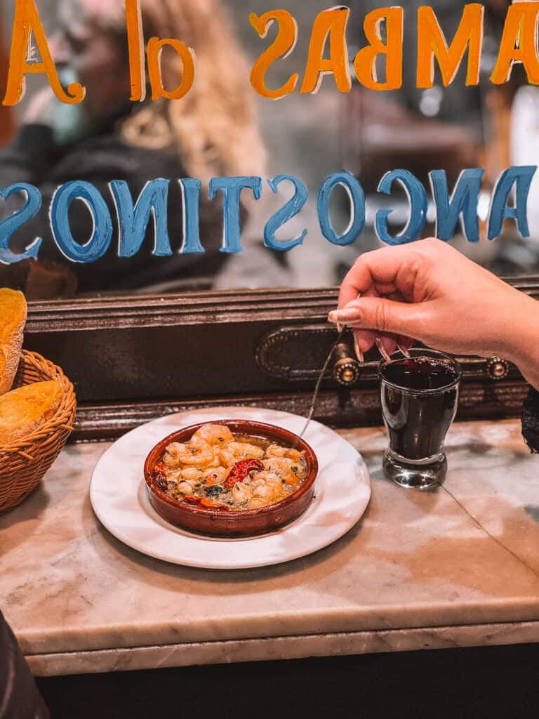 A customer's point of view at a Spanish restaurant, holding a fork over a sizzling clay dish of garlic shrimp, with a glass of red wine and a basket of bread in the background.