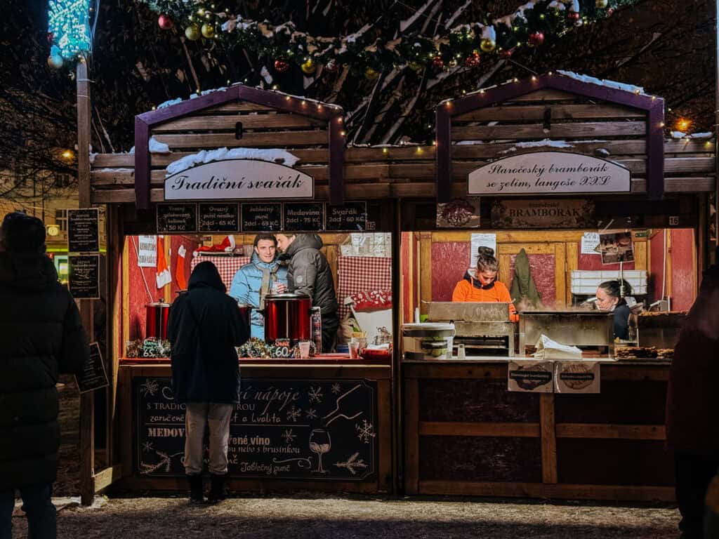 An evening scene at a Christmas market where vendors serve steaming beverages from a traditional "svarak" stall adorned with Christmas lights and snow.