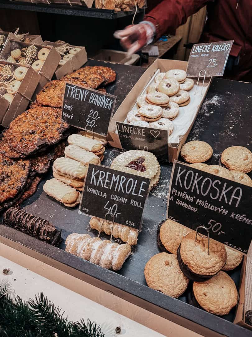 Delectable Christmas pastries on display at a market stall, including "kremrole" (cream rolls) and "kokoska" (coconut pastries), with prices labeled in Czech koruna.