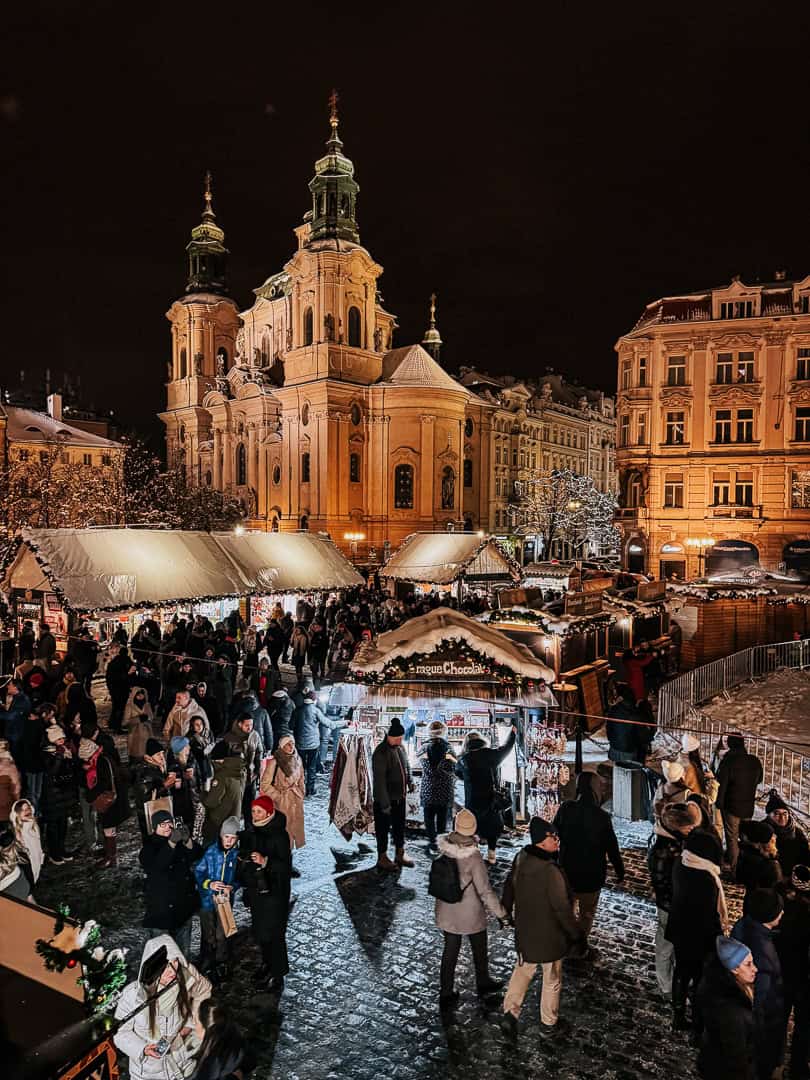 Snow-covered roofs of Christmas market stalls with crowds of visitors enjoying the festive atmosphere near the Baroque Saint Nicholas Church in Prague