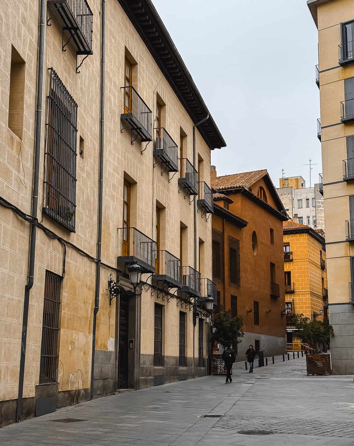 A quiet street scene with traditional European architecture, featuring buildings with textured facades and wrought-iron balconies, under a cloudy sky