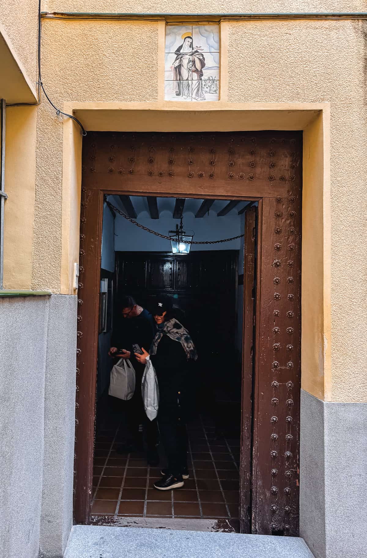 Two people exiting through a large wooden door under a framed religious tile painting, with one person looking at a mobile device