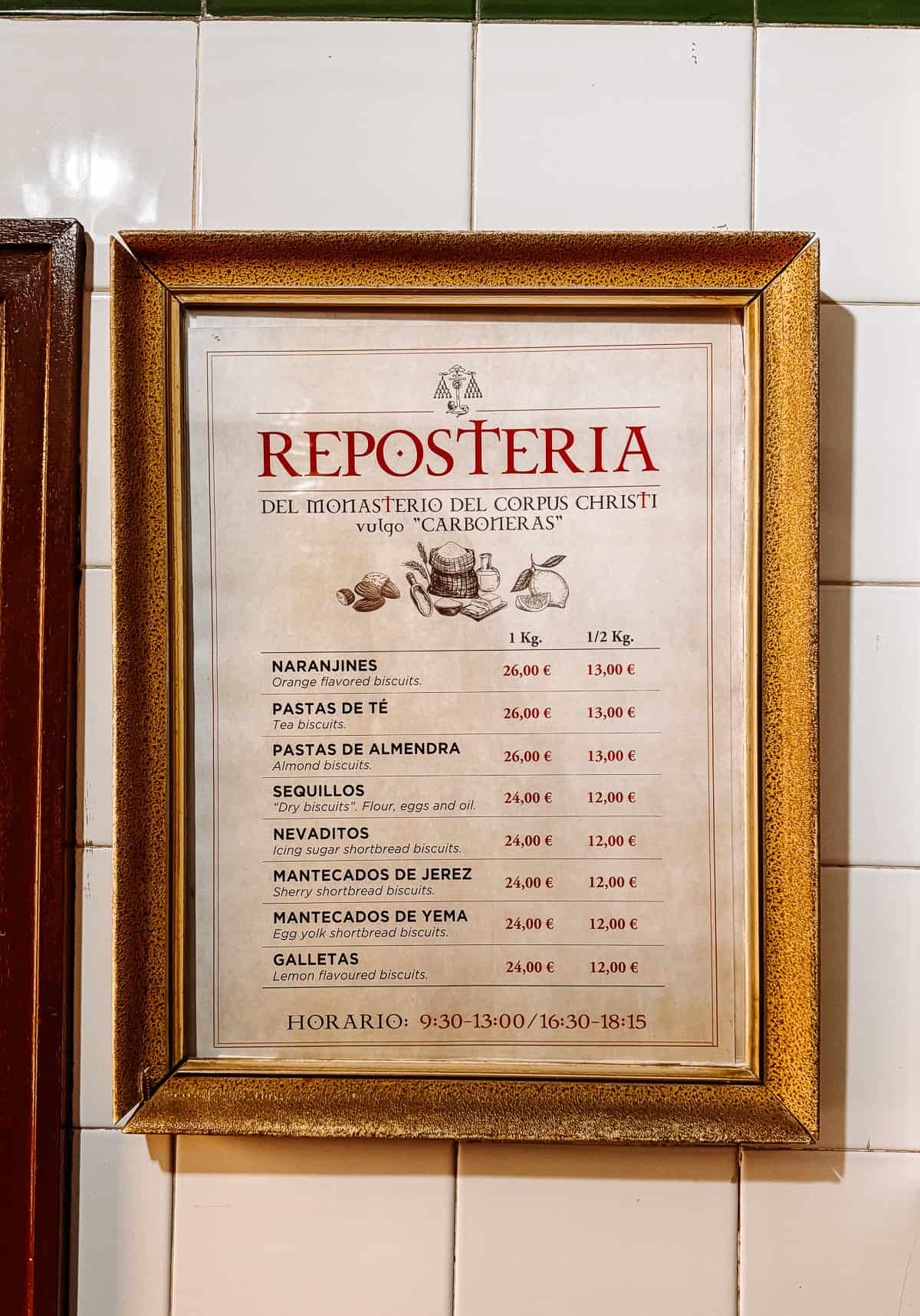 Framed price list on a tiled wall for 'REPOSTERIA DEL MONASTERIO DEL CORPUS CHRISTI vulgo "CARBONERAS"' showing various traditional Spanish biscuits and their prices, under the 'HORARIO' for sales and visits