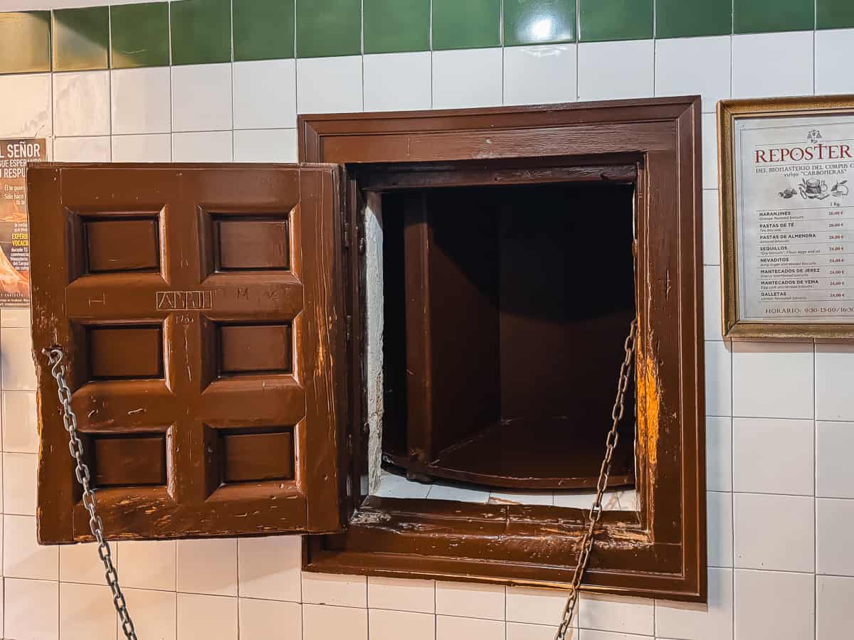 n open turnstile window in a tiled wall with a heavy brown wooden door, part of a traditional Spanish monastery selling sweets