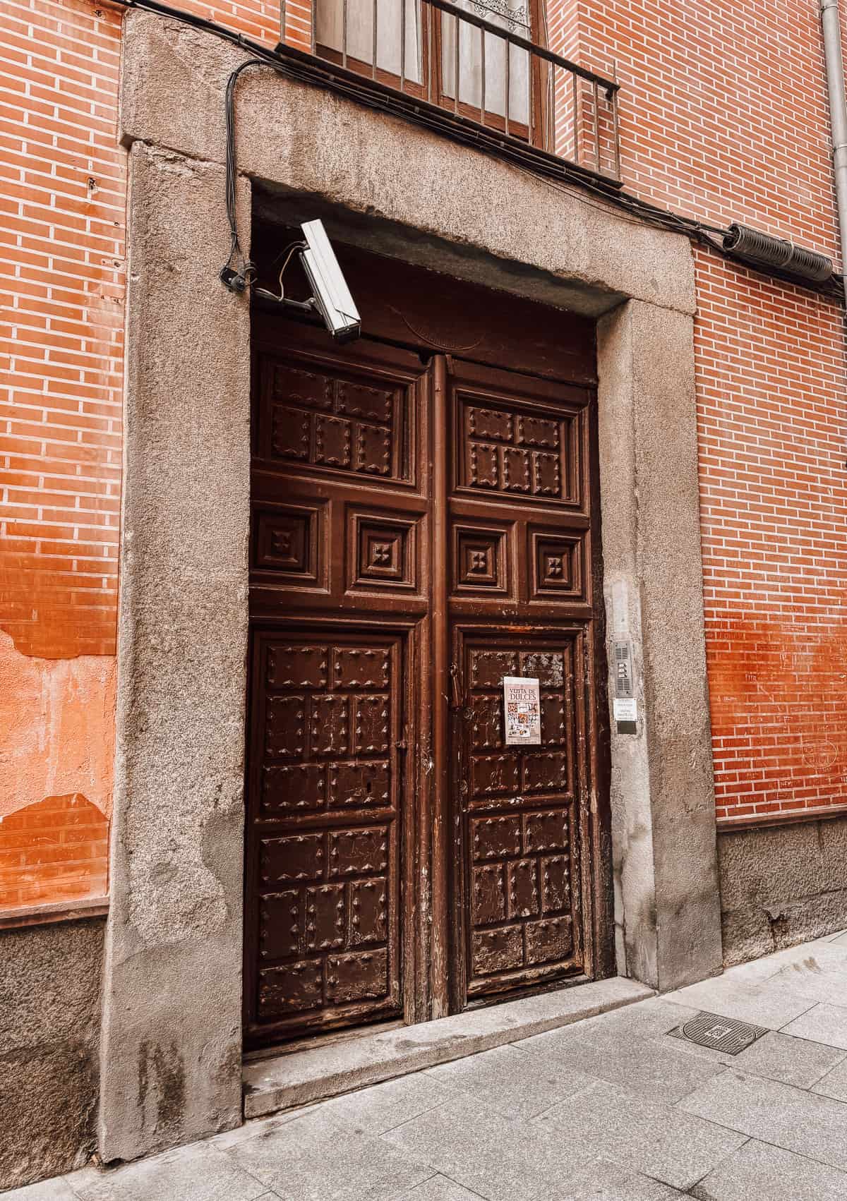 Old ornate wooden double doors with a 'Venta de Dulces' flyer and intercom buzzer, set in a red brick wall of a historic Spanish building