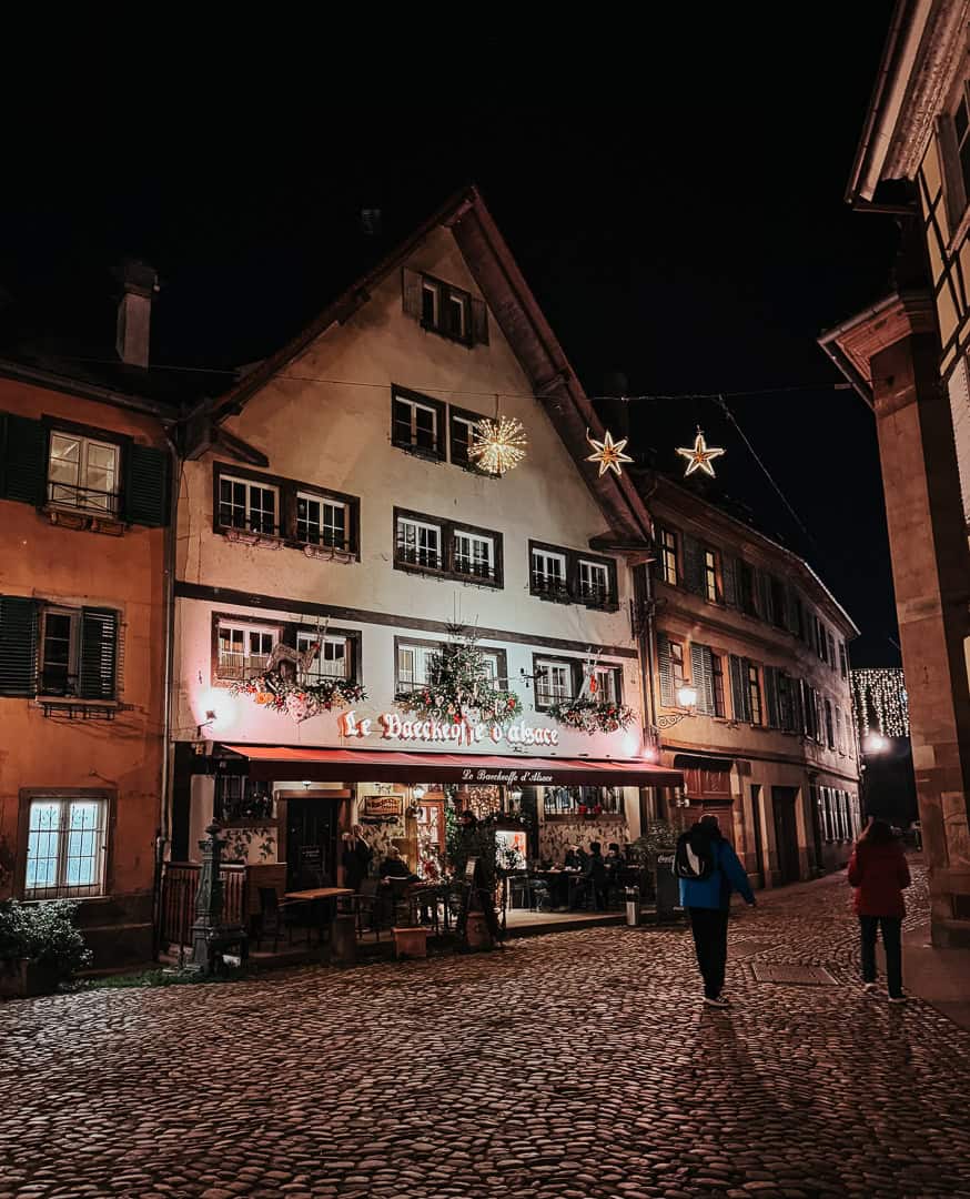 The warm glow of Christmas illuminates "Le Baeckeoffe d'Alsace" in Strasbourg, with star-shaped lights strung across the street and people walking on the cobblestones.