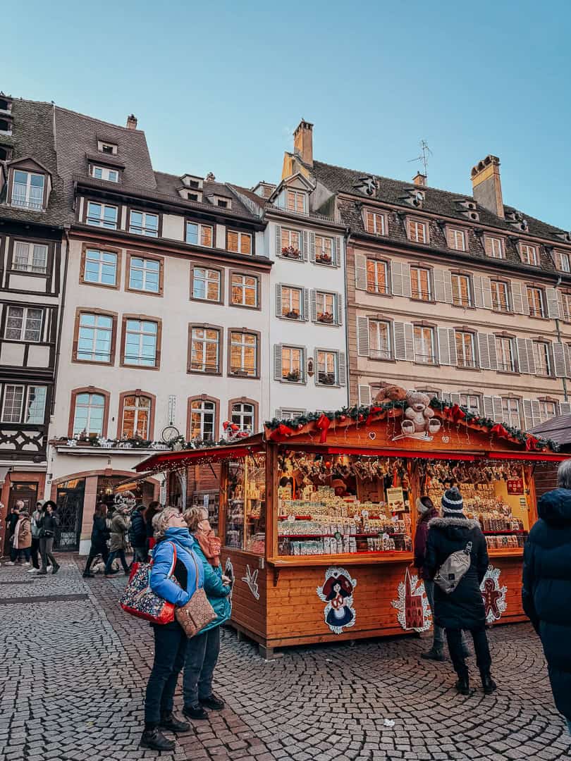 A well-lit Christmas market booth nestled under a tree with sparkling ornaments, offering an array of colorful toys and gifts to evening shoppers.