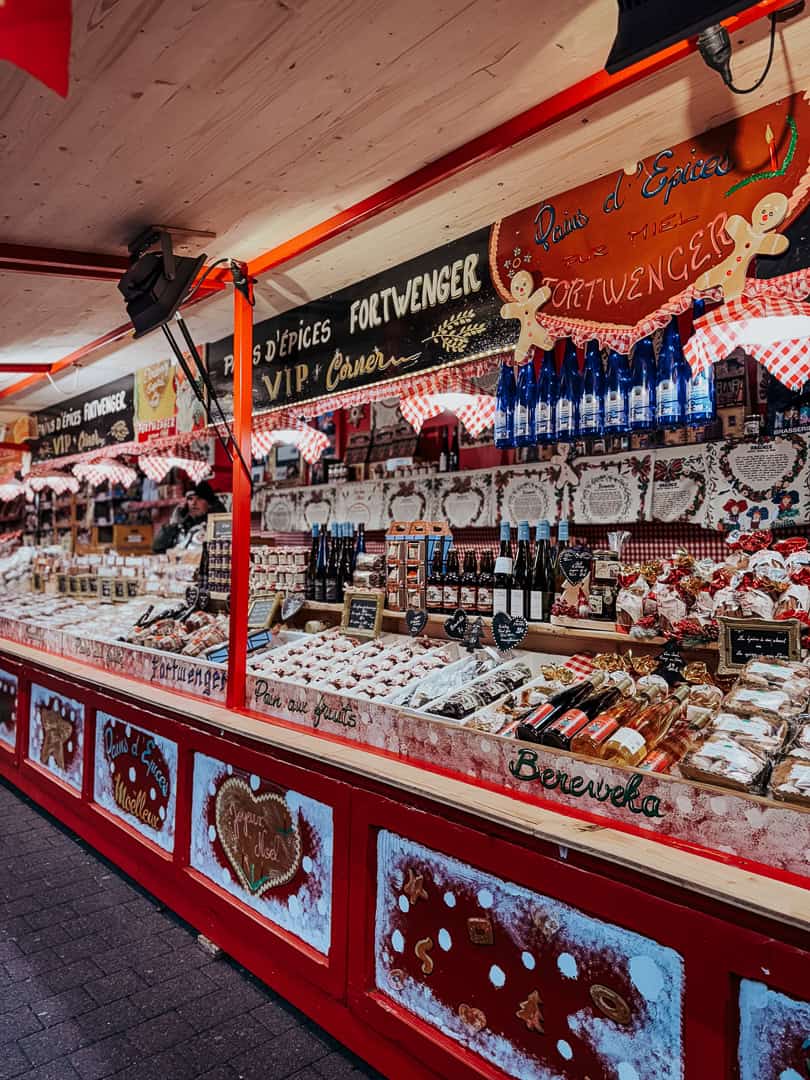 A festive market stall in Strasbourg is filled with gingerbread and assorted regional delicacies under a sign reading "Pains d'Épices FORTWENGER," all adorned with Christmas decorations.