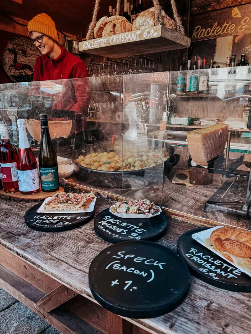 Cheerful market stall offering raclette and croissants, with a clear view of the melting cheese and bottles of wine