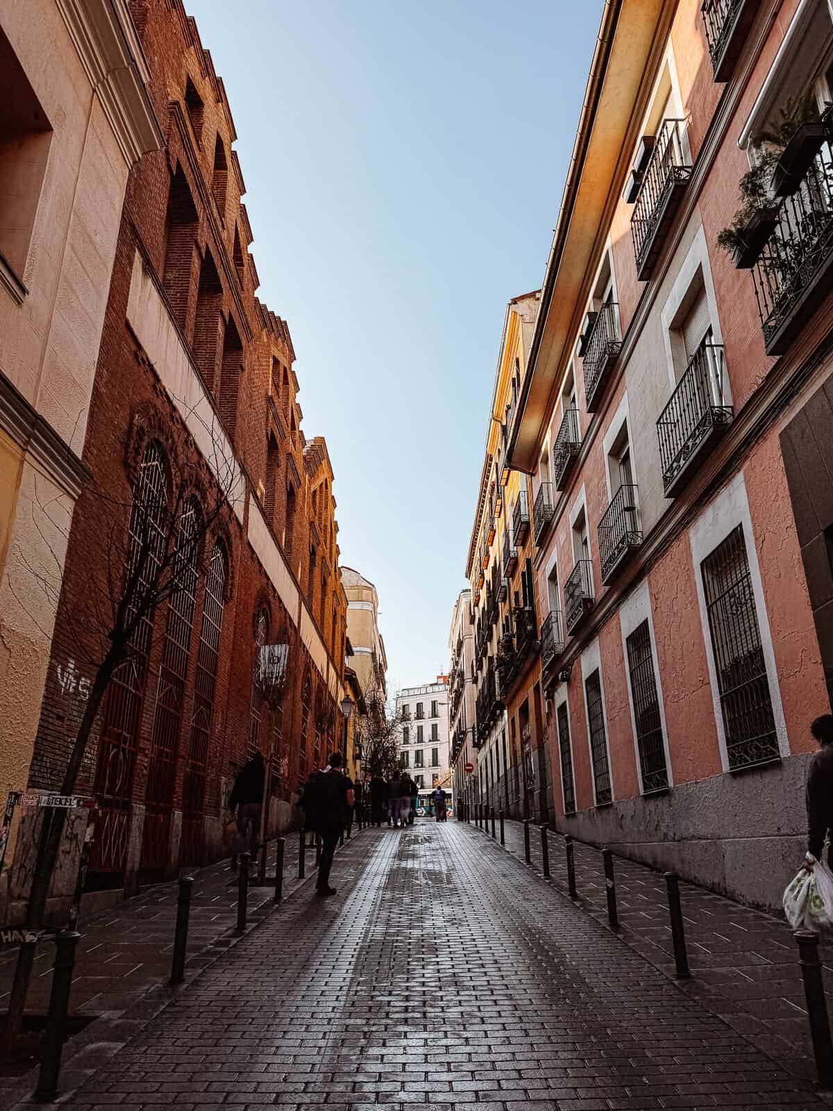 A serene cityscape depicting a cobblestoned street flanked by traditional European buildings under a clear blue sky in Madrid, Spain.