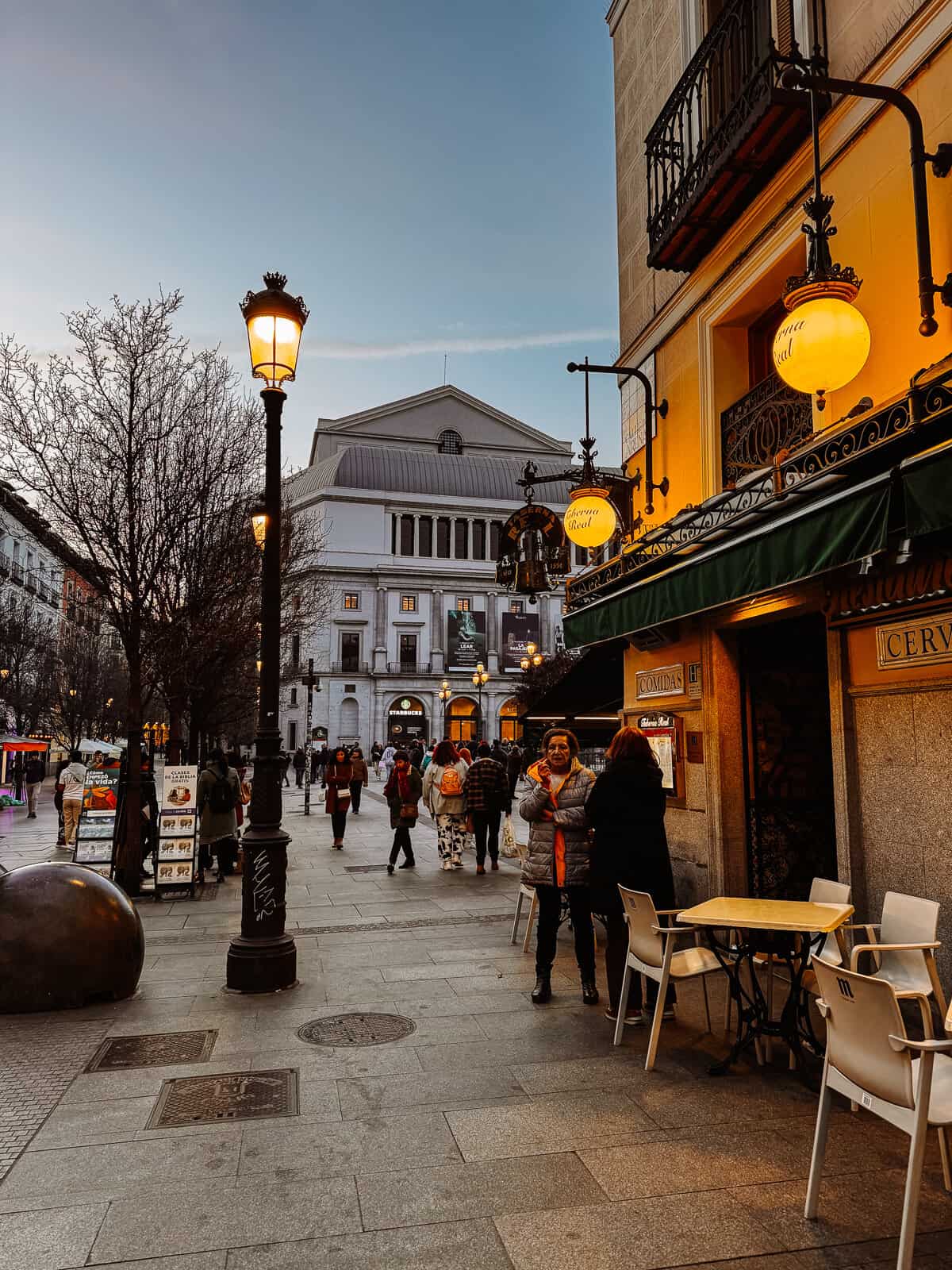 A lively street scene at dusk in Madrid, highlighting warm glowing street lamps, an array of shops, and people gathering outside a cafe.