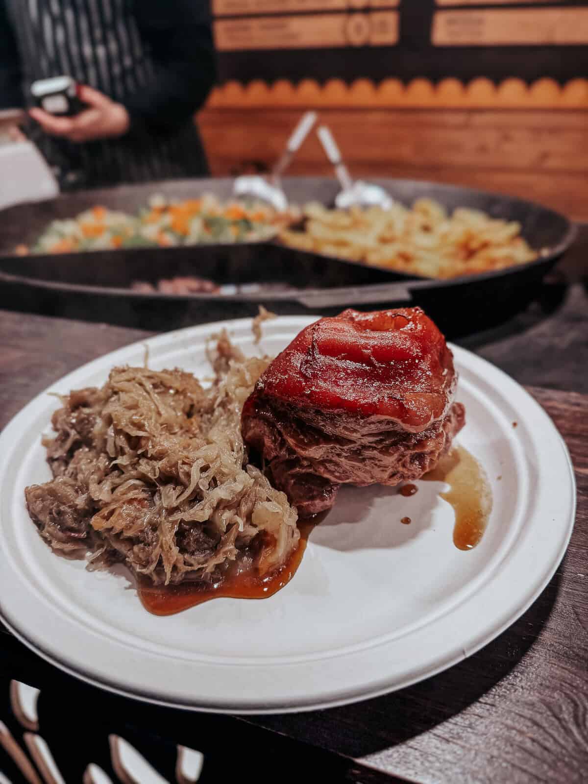 A close-up of a hearty meal featuring a roasted pork knuckle glazed with a shiny sauce, accompanied by sauerkraut, served on a white plate at a market stand