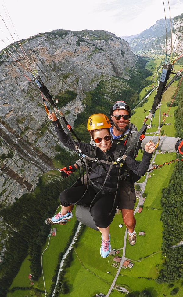 Thrilled duo paragliding over a lush green valley in Switzerland, with the woman in the forefront giving a thumbs-up and both wearing helmets and secure harnesses, against a dramatic backdrop of towering cliffs and a picturesque rural landscape far below.