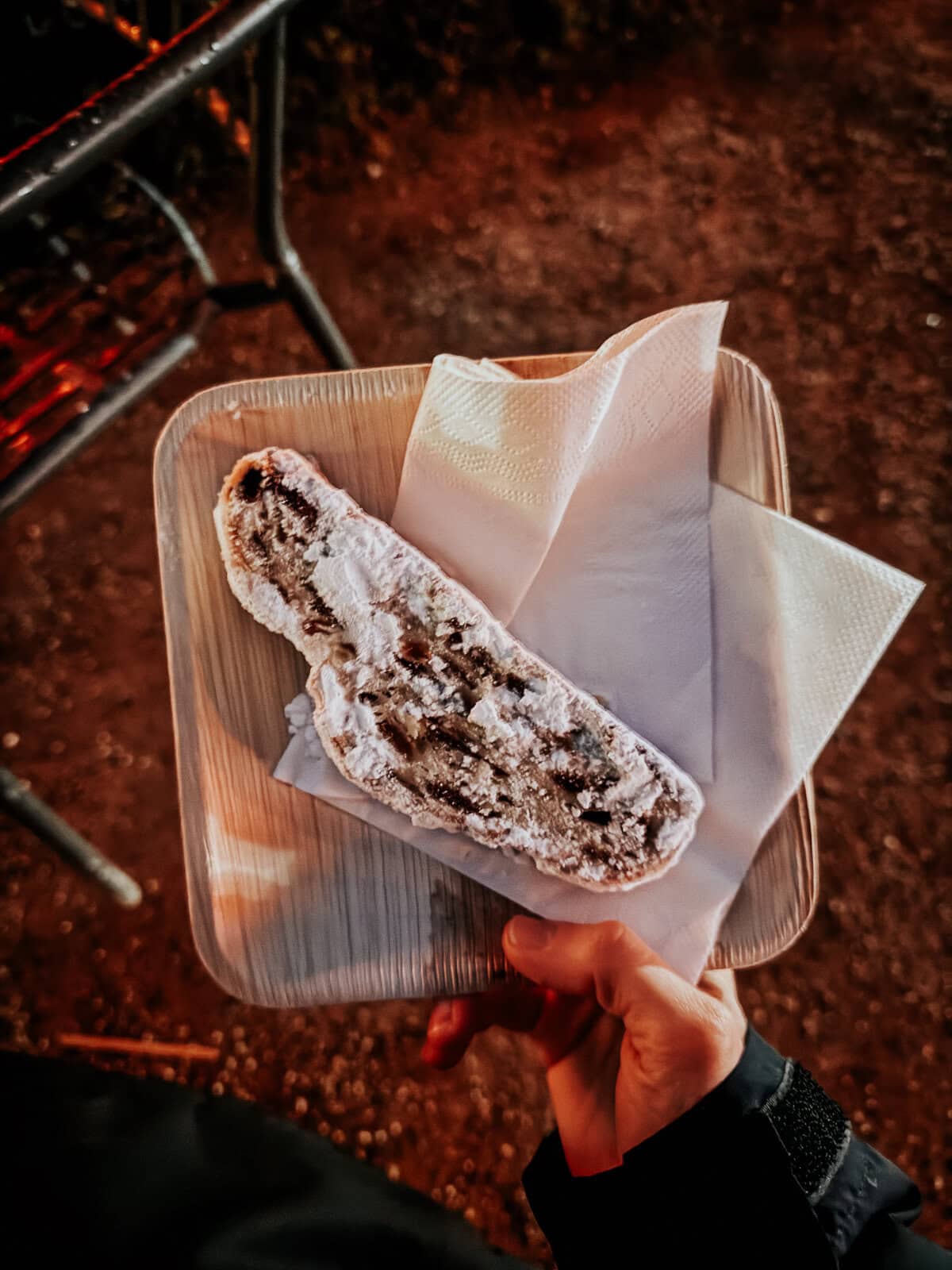 A hand holding a slice of stollen, a traditional German fruit bread, dusted with powdered sugar, with a backdrop of festive lights.