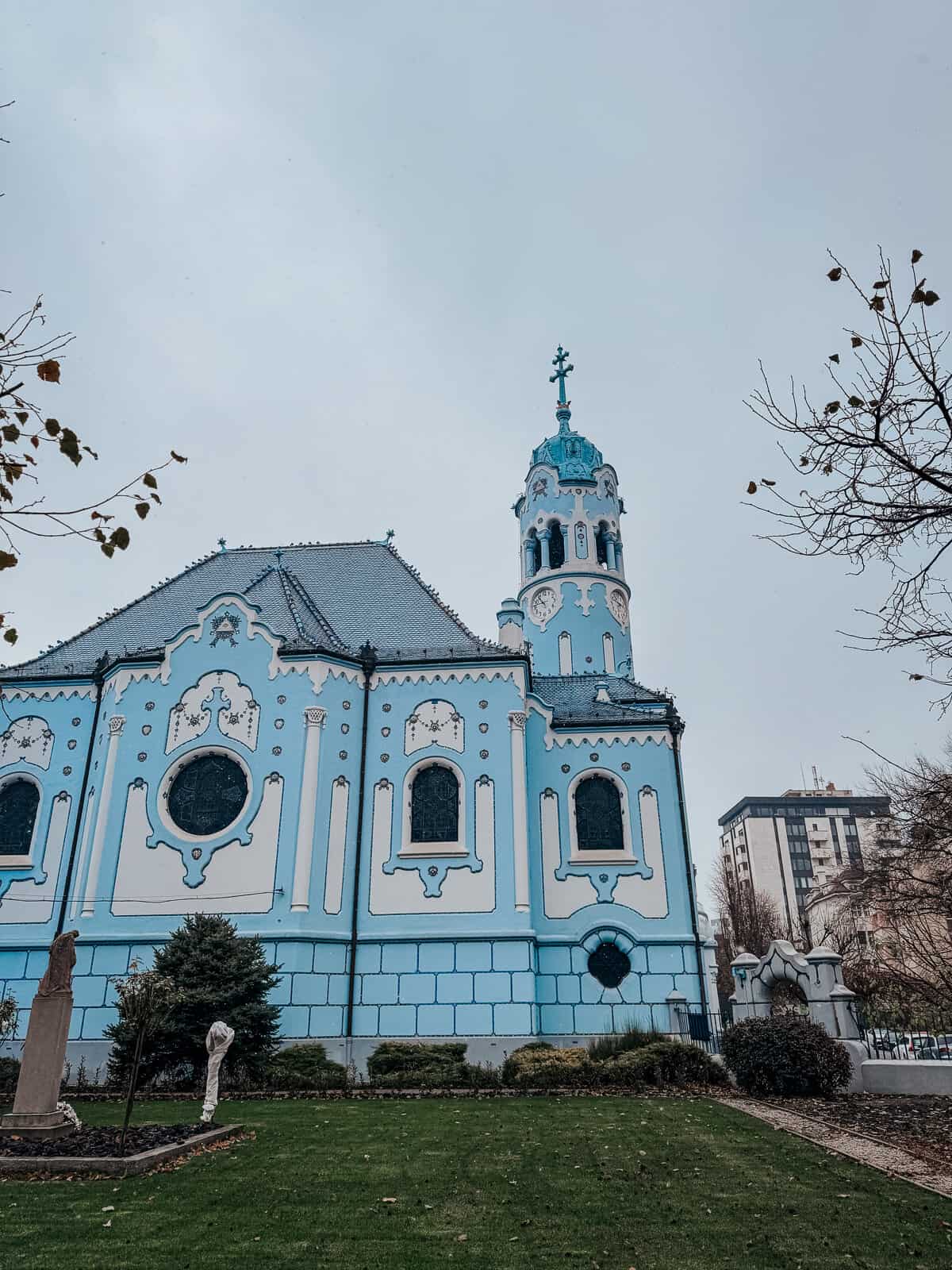 A vibrant blue church with ornate white detailing stands against a grey sky, with bare trees and a green lawn in the foreground.
