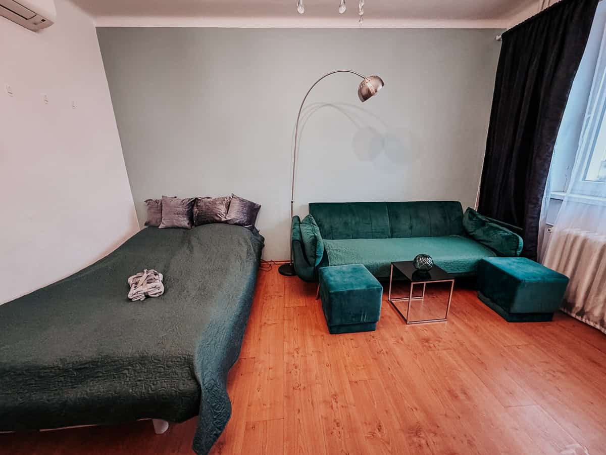 A simple but cozy room featuring a single bed and a green velvet sofa set against soft green walls, minimal decor, and natural light.