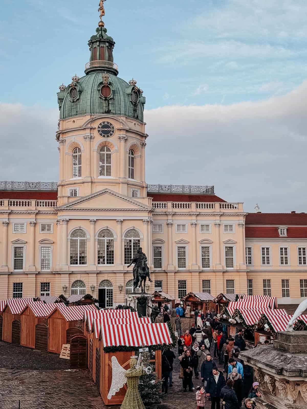 A bustling Christmas market in the courtyard of an ornate palace with a green dome. Stalls with red and white striped canopies are surrounded by holiday decorations and visitors in winter attire