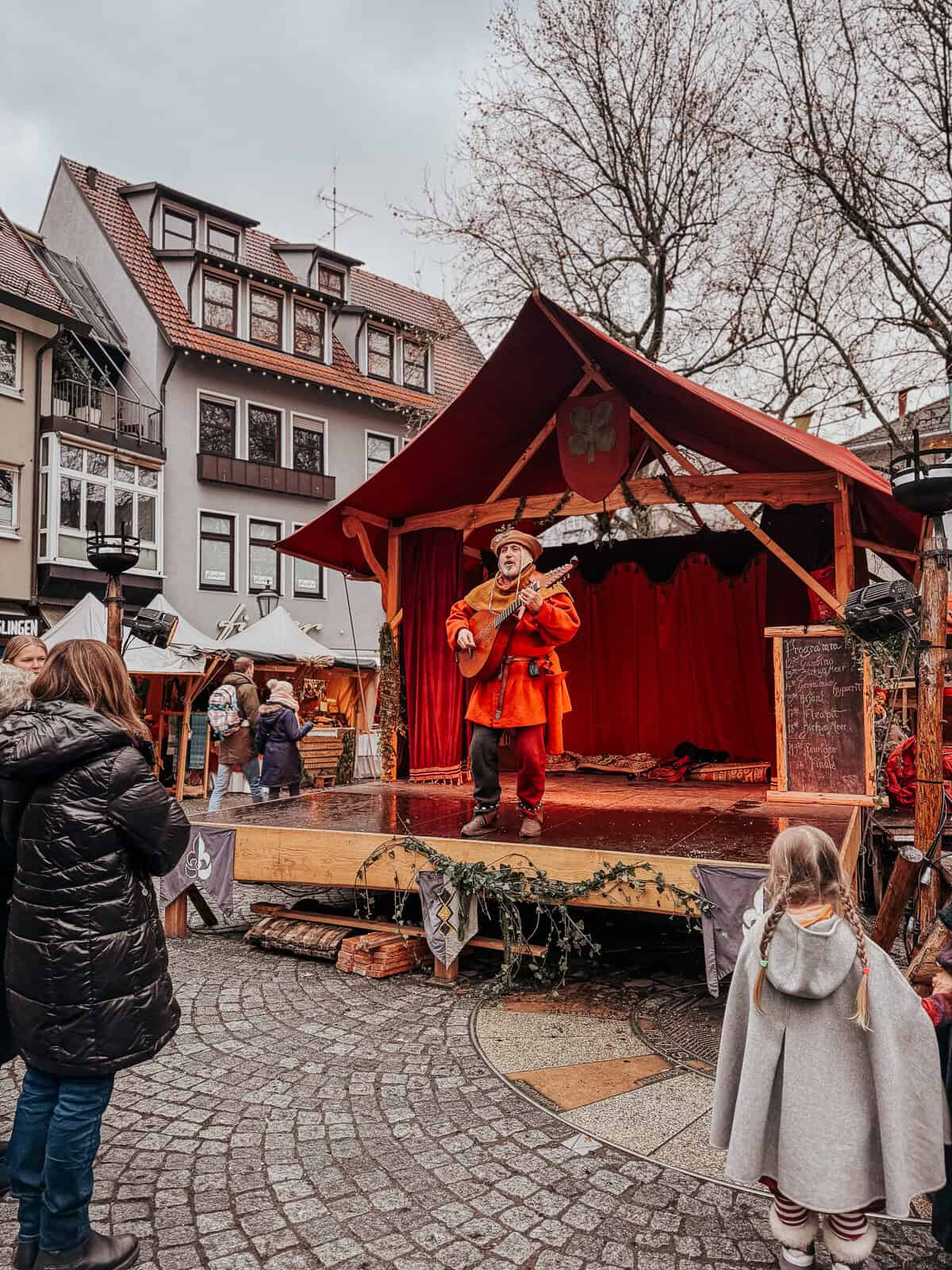a minstrel performing on a stage in medieval attire, entertaining children and adults gathered around.