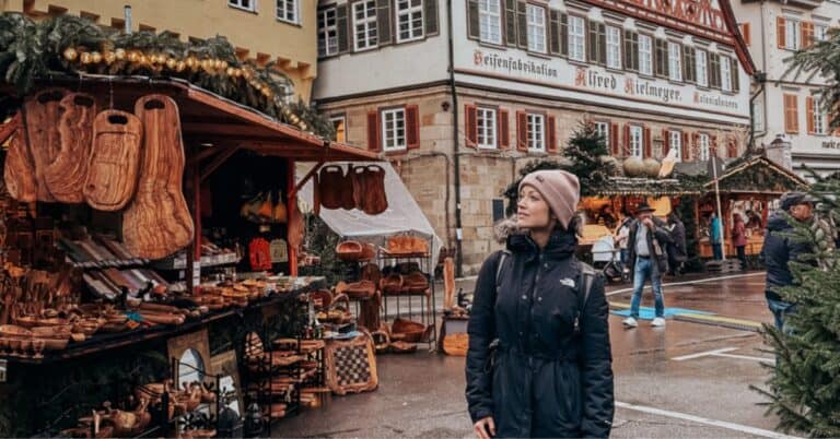 A person stands contemplatively in front of a traditional German Christmas market, with wooden stalls selling handcrafted goods, against the backdrop of a striking half-timbered building in Esslingen