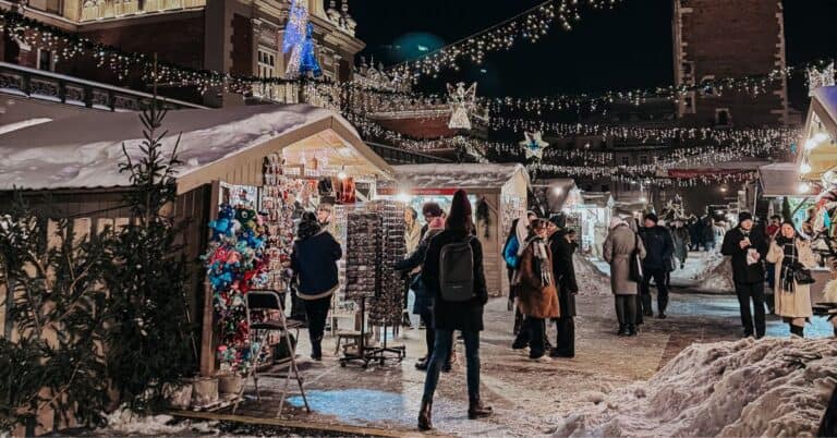 Night scene at a Christmas market with a view of the historic cloth hall illuminated by festive lights, snowy paths, and shoppers browsing through various stalls