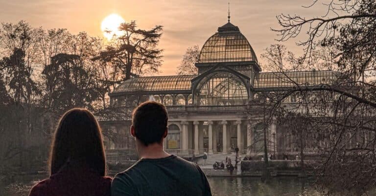 a couple standing with their backs to the camera with the mans arm around the woman. They are standing in front of the crystal palace at sunset