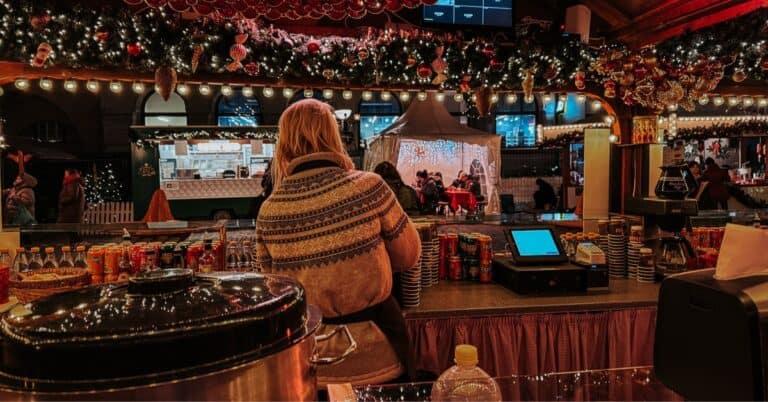 View from a market vendor's stall showing a large pot, beverages, and the back of a vendor looking out at the busy Christmas market decorated with red Christmas balls and lights.