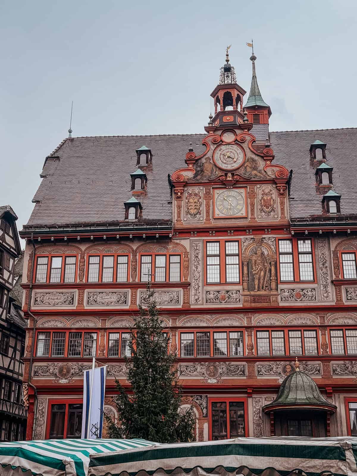 The ornate facade of a richly decorated building in Tübingen, featuring intricate frescoes and a historic clock, with a Christmas tree in front.