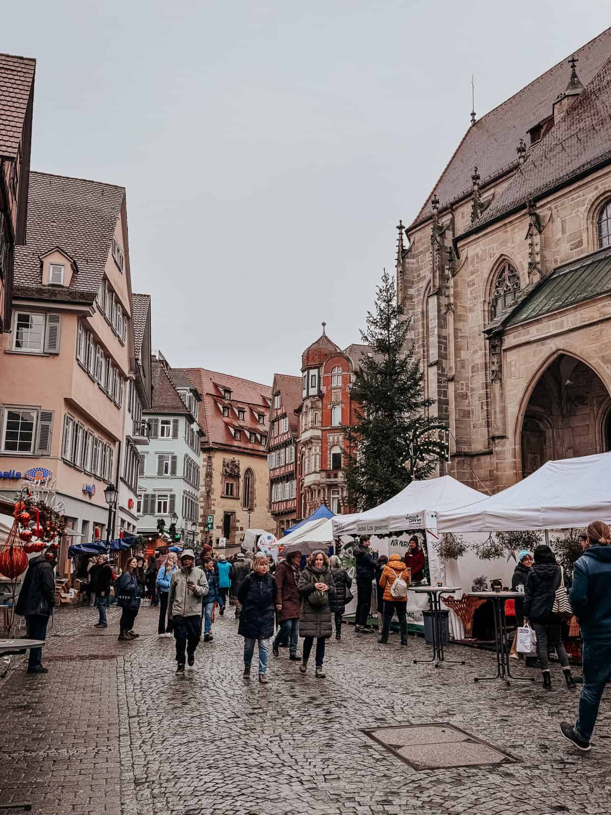 View of a busy street market adjacent to a large Gothic church in Tübingen, with people browsing stalls under overcast skies."