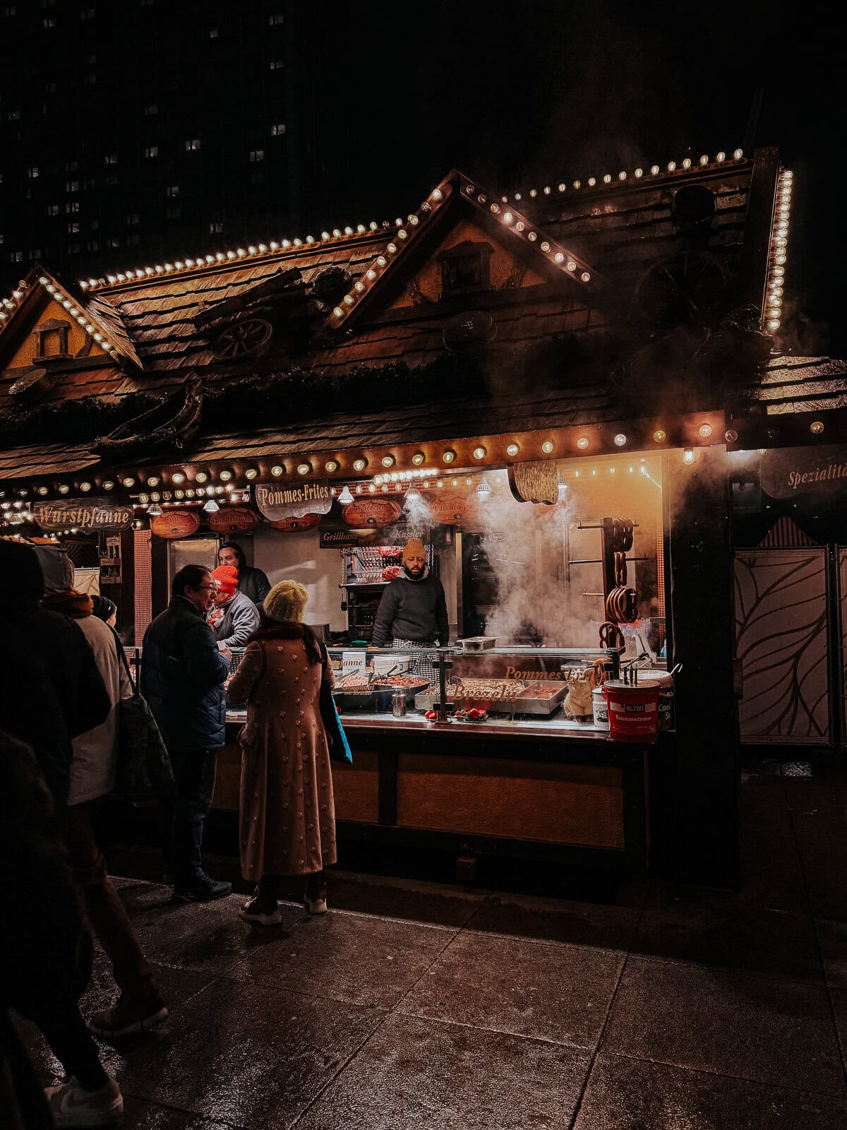 A bustling outdoor food stall at night, decorated with festive lights and emitting steam from cooking. People are gathered around, ordering and waiting for food, creating a lively winter market scene