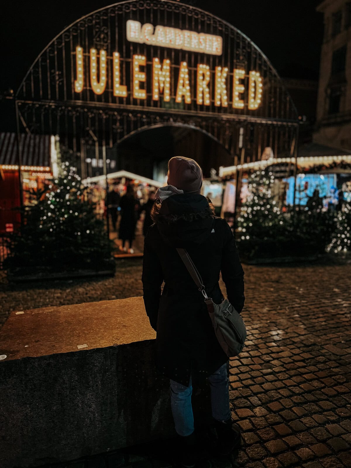 A woman viewed from behind, looking at a brightly lit archway sign reading "HC Andersen Julemarked" (HC Andersen Christmas Market), with festive lights and decorations around.